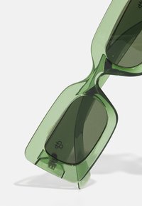 Roheline prillid, millel on massiivne geomeetriline disain, läbipaistev plastraam ja tumedad läätsed. Silmapaistev kumer detail sillal.