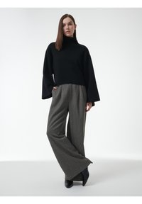 Vrouw in een zwart coltrui en wijde grijze textuur broek, staand met één hand in de zak tegen een plain witte achtergrond.