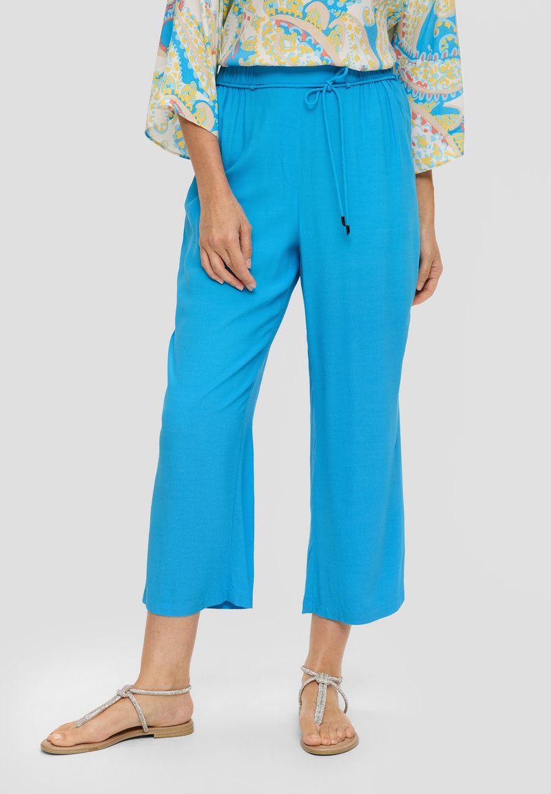 Femme portant un pantalon large bleu vif coupé au-dessus de la cheville avec une taille à cordon et des sandales plates argentées à brides, debout devant un fond gris.