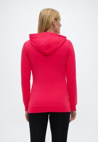 Vaaleatukkainen nainen, joka pukeutunut kirkkaanpunaiseen huppariin ja mustiin housuihin, kuvattuna takaapäin yksinkertaista valkoista taustaa vasten.