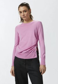 Maglione rosa a maniche lunghe con scollatura rotonda, bottoni laterali e dettagli arricciati in vita. Abbinato a pantaloni neri.