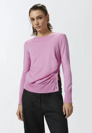 Maglione rosa a maniche lunghe con scollatura rotonda, bottoni laterali e dettagli arricciati in vita. Abbinato a pantaloni neri.