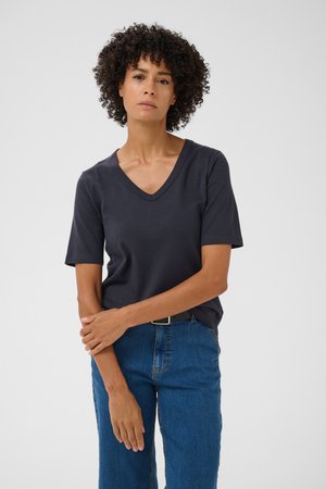 Une femme aux cheveux foncés et bouclés porte un tee-shirt foncé à col en V et un jean bleu sur un fond uni et clair.