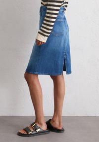 Denim A-lijn rok in medium blauw, middenlang met een rechte zoom en een subtiele split aan de achterkant. Geddraagd met zwarte sandalen met een gouden gesp.