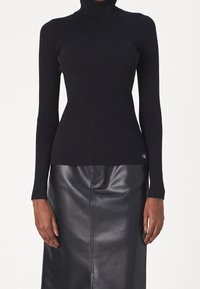 Kvinna som bär en åtsittande svart ribbad polotröja och en högmidjad svart läderkjol, stående med armarna avslappnat ned längs sidorna.