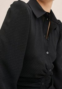 Schwarze Button-Up-Bluse aus strukturiertem Stoff mit gepunktetem Muster, einem Kragen und gerafftem Detail an der Taille.