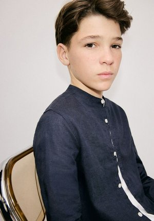 Tinejdžer sa kratkom smeđom kosom, odjeven u tamnu košulju na kopčanje, sjedi na stolici i ozbiljno gleda prema kameri.