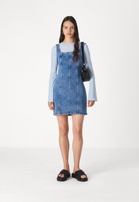 Rochie din denim albastră, cu bretele ajustabile, buzunare laterale și detalii de coasere vizibile. Asortată cu un top cu mânecă lungă de un albastru deschis.