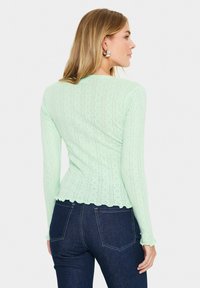 Haut à manches longues vert menthe avec un motif tricoté texturé, un ourlet en dentelle et une silhouette ajustée. Associé à un jean en denim foncé.