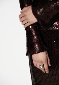 Bordeaux pailletten jurk met uitlopende mouwen, voorzien van een golvende zoom en glanzende textuur. Zilveren ring siert de hand.