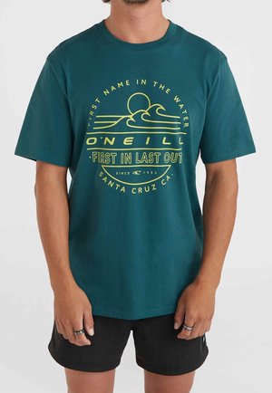 Teal Baumwoll-T-Shirt mit einem gelben grafischen Design mit Wellen und Text. Kurze Ärmel und eine lockere Passform, kombiniert mit schwarzen Shorts.