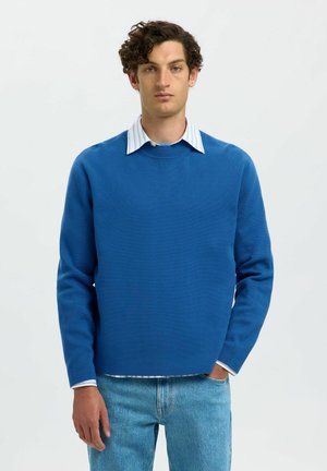 Young man with curly hair wearing a blue crewneck sweater over a white striped collared shirt and light blue jeans, standing against white background.