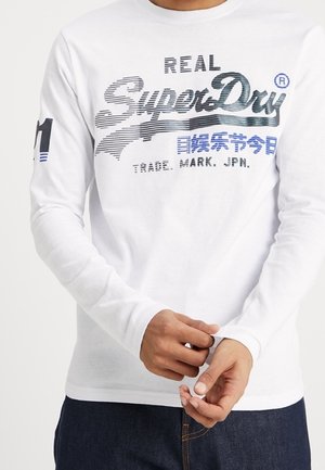 Weißes Langarm-T-Shirt mit einem blauen und schwarzen grafischen Print-Logo und Text auf der Vorderseite. Weicher Baumwollstoff mit Rundhalsausschnitt.