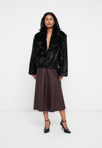 Giacca in pelliccia sintetica nera con ampio colletto, abbinata a una gonna in pelle bordeaux lunghezza ginocchio. La modella indossa scarpe nere con tacco alto.