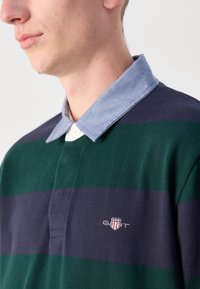 Polo tričko s zeleno-námornickými pruhy, které má límec z chambray a malé vyšívané logo na hrudi. Materiál vypadá měkce a je strukturovaný.