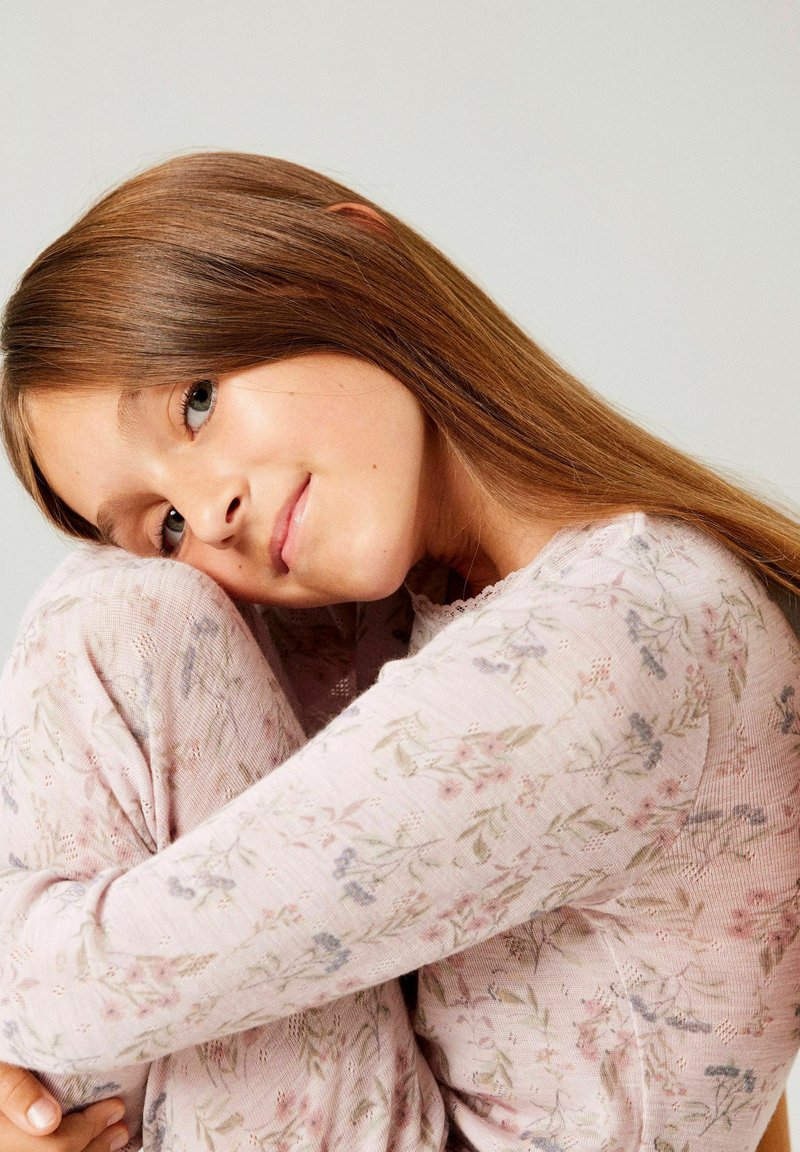 Jeune fille aux longs cheveux bruns portant un pyjama floral rose clair, assise avec les genoux serrés, souriant doucement sur un fond uni.