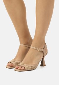 Sandalias de tacón alto beige con un diseño de textura trenzada, punta abierta y correa en el tobillo con hebilla dorada. Altura del tacón de 3 pulgadas.