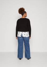 Maglietta nera a maniche lunghe con un pannello posteriore bianco a pois; abbinata a jeans a gamba larga blu. La maglietta ha un collo rotondo e un dettaglio a spacco.
