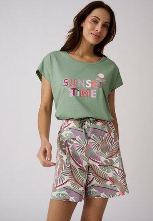 Mujer con una camiseta verde con el texto "SUNSET TIME" y pantalones cortos con estampado floral atados a la cintura sobre un fondo liso.