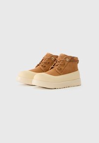 Bottines en daim tan avec semelles et lacets en caoutchouc beige, arborant le logo UGG sur la languette, présentées sur un fond blanc uni.