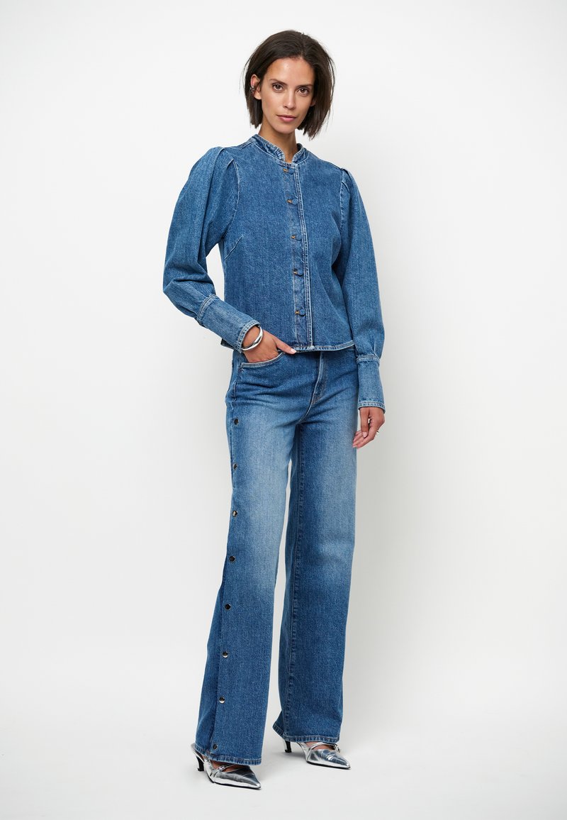 Denim jack met een hoge kraag en knopen aan de voorkant, gecombineerd met uitlopende denimbroeken met knopen aan de zijkant. Beide zijn in middelblauw.