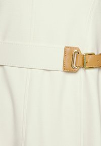 Krémová látka s hladkým povrchem, která má široký, elastický pás z hnědé kůže se zlatým kováním.