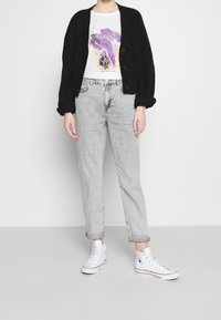 Svart stickad kofta med stora knappar, vit t-shirt med lila och guldmönster, ljusgrå raka jeans och vita sneakers.