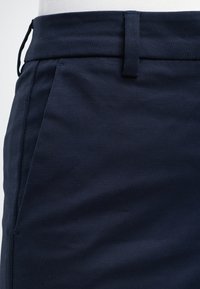 Gros plan d'une personne portant un pantalon bleu marine foncé avec une ceinture, des passants de ceinture et une poche latérale, associé à un haut blanc.