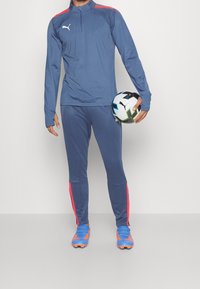 Mies sinisessä Puma-verryttelypuvussa pitelee Puma-jalkapalloa, jalassaan siniset ja oranssit nappikset, seisoo yksinkertaisen valkoisen taustan edessä.