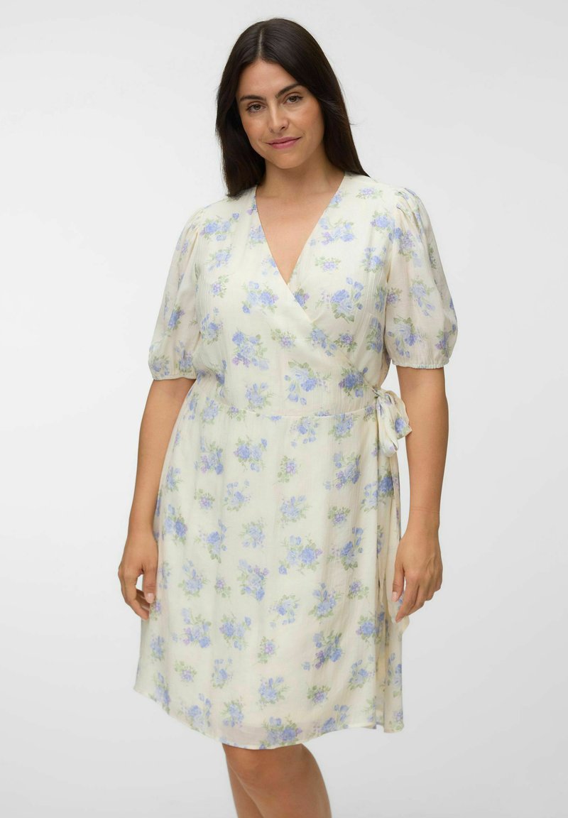 Femme portant une robe portefeuille crème mi-longue avec imprimé floral bleu et manches courtes bouffantes, debout devant un fond clair uni.