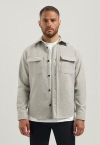 Man in een lichtgrijze button-up jas over een wit shirt, staand tegen een effen achtergrond, met het gezicht naar voren en een neutrale uitdrukking.