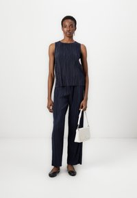 Haut sans manches plissé bleu marine associé à un pantalon large assorti. Le mannequin tient un petit sac à main blanc et porte des ballerines noires. Fond minimal.