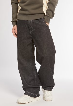 Pantaloni larghi in denim nero con cuciture bianche, tasche frontali e vestibilità rilassata, abbinati a sneakers di colore chiaro.