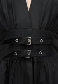 Gros plan sur un vêtement noir avec deux sangles horizontales munies de boucles en argent et d'œillets sur un tissu froncé.