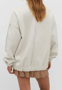 Felpa oversize grigia chiara con spalle cadenti ed orlo elastico. Tessuto morbido e design della schiena senza cuciture; abbinata a una gonna beige plissettata.