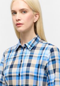 Blå og beige ternet skjorte med knapper. Har en klassisk krave og en tætsiddende pasform med varierende firkantede mønstre og mørke knapper.
