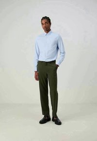 Camisa de botão azul claro, tecido leve; calças verdes escuras ajustadas; sapatos pretos com design recortado; pose de pé.
