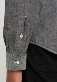 Polsino di camicia in lino grigio con pattina e bottoni bianchi. Il tessuto ha una lavorazione a trama e il polsino è leggermente arricciato.