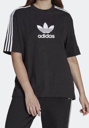 Person trägt ein schwarzes Adidas-T-Shirt mit weißem Logo und drei Streifen auf den Ärmeln, kombiniert mit schwarzen Hosen mit weißen Streifen.