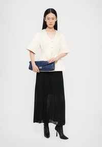 Krēmkrāsas, īsām piedurknēm tops ar pogām; melna plisēta maksi svārki; zils krokodila tekstūras kabatas. Modelis valkā melnus bata-nodaudprāvas zābakus.