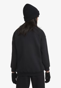 Sweat-shirt noir à coupe décontractée, avec des poignets et un ourlet côtelés. Associé à un bonnet en tricot noir et des gants noirs, porté comme une tenue complète.