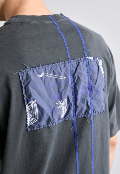 Black t-shirt featuring a fabric patch with blue graphics, textured finish, and stitched detailing. Visible fraying around the patch edges.