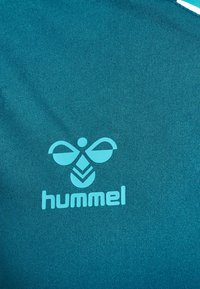 Tissu bleu sarcelle avec un logo hummel bleu clair centré représentant une abeille stylisée au-dessus du nom de la marque en minuscules.