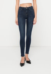 Jeans oscuros de tiro alto y corte ajustado, con cinco bolsillos, pasadores para cinturón y una textura desgastada. Combinados con zapatos negros de punta.