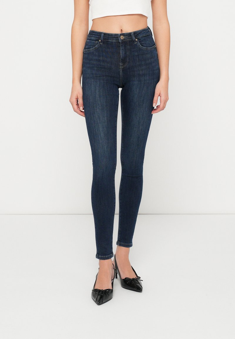 Jeans oscuros de tiro alto y corte ajustado, con cinco bolsillos, pasadores para cinturón y una textura desgastada. Combinados con zapatos negros de punta.