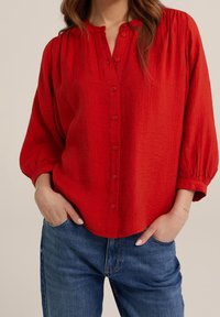 Red button-up shirt with a textured fabric, relaxed fit, and puffed sleeves, paired with blue denim jeans.