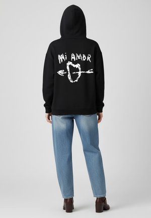 Zwarte hoodie met witte grafische tekst "Mi Amor" en een hart met pijl. Gecombineerd met lichtblauwe, high-waisted spijkerbroeken en bruine hakboots.
