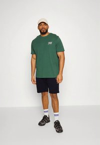 Green cotton t-shirt with logo, paired with black shorts and black sneakers. White socks with blue stripes are visible. Model stands upright.