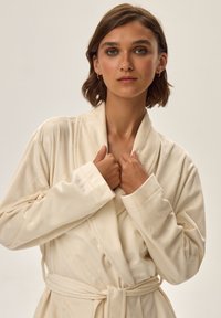 Kremowy szlafrok z miękkiego, teksturowanego materiału. Posiada kołnierz szalowy, długie rękawy i pasek w talii. Widoczny subtelny wzór geometryczny.