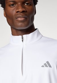 Biely športový zipsový top vyrobený z hladkej tkaniny; má vysoký golier, sivé logo a kovový zips.